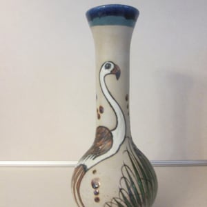May include: A ceramic vase with a white background and a painted design of a flamingo in brown, white, and blue. The vase has a round base and a long, narrow neck.