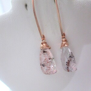 May include: A pair of dangle earrings with copper-colored ear wires. Each earring features a clear, teardrop-shaped stone with red and black inclusions, wrapped in copper wire. The earrings are displayed on a white jewelry stand.