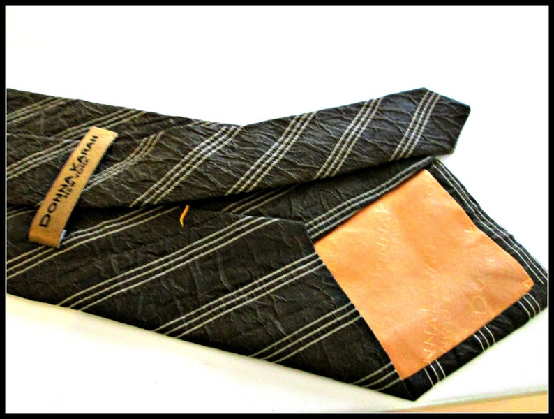 May include: A black and white striped tie with a brown textured fabric. The tie has a brown leather label with the text "DONNA KARAN NEW YORK" printed on it.