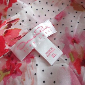 May include: Close-up of a floral print fabric with a white label. The fabric features red and pink flowers on a white background with black polka dots. The label reads "Oscar de la Renta" and "Made in Cambodia XL".