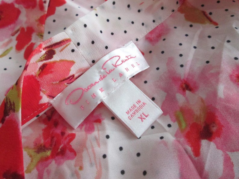 May include: Close-up of a floral print fabric with a white label. The fabric features red and pink flowers on a white background with black polka dots. The label reads "Oscar de la Renta" and "Made in Cambodia XL".