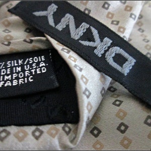 May include: A beige silk tie with a repeating pattern of small brown squares. The tie has a black label with the text "D'ANN" and the words "100% SILK/SOIE MADE IN U.S.A. OF IMPORTED FABRIC".