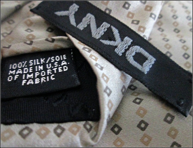 May include: A beige silk tie with a repeating pattern of small brown squares. The tie has a black label with the text "D'ANN" and the words "100% SILK/SOIE MADE IN U.S.A. OF IMPORTED FABRIC".