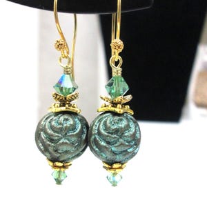 May include: A pair of dangle earrings with gold-colored hooks and accents. Each earring features a teal-colored, rose-patterned bead, a green crystal, and a smaller green crystal at the bottom. The earrings are displayed against a black background.