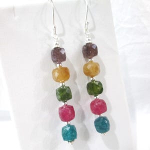 May include: A pair of dangle earrings featuring five faceted, multi-colored gemstone beads arranged in a vertical line. The beads are purple, yellow, green, pink, and blue, separated by small silver-toned spacers and attached to silver-toned hooks.