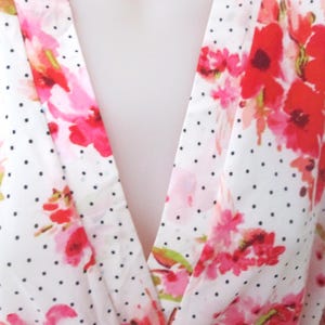 May include: White fabric with a pattern of red and pink floral designs and small black polka dots. The fabric is draped, creating a V-neck shape. The floral pattern is a watercolor style.