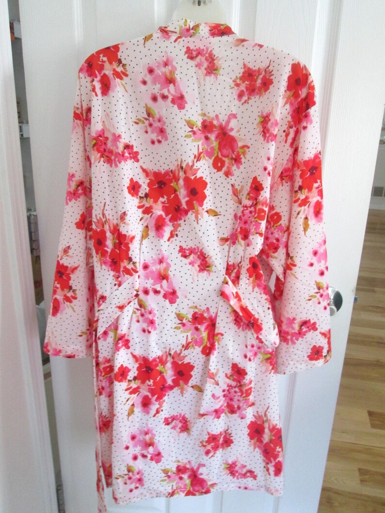 May include: A white robe with a floral pattern featuring red and pink flowers and small black polka dots. The robe has long sleeves and a tie belt. The floral pattern is repeated throughout the garment.