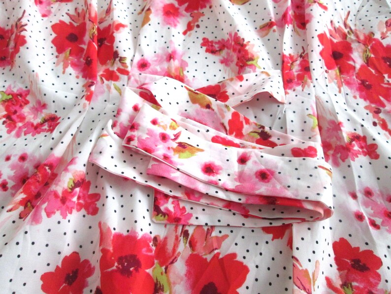 May include: White fabric with a pattern of red and pink flowers and small black polka dots. The fabric is folded, revealing a cuff with the same floral and polka dot design. The overall theme is floral.