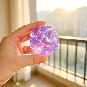 May include: A translucent, spherical object with a floral design. The object is a mix of purple and pink hues, with a glossy finish. The background shows a balcony with a metal railing and a cityscape in the distance.