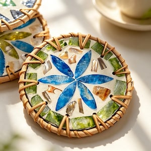 May include: Round coaster with a mosaic design featuring a blue flower and accents of green, tan, and clear glass. The mosaic is set on a cream-colored background and framed by a woven rattan border.
