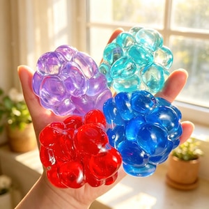 May include: Four colorful, translucent stress balls in shades of purple, teal, blue, and red. Each ball is composed of numerous rounded shapes, creating a textured, bubbly appearance. The balls are held in a hand, with natural light illuminating them.