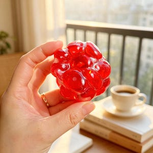 May include: A red, translucent stress ball held in a hand. The ball has a bumpy, textured surface and a rounded shape. The background includes a cup of coffee, books, and a window.