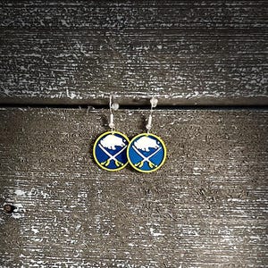 May include: A pair of round earrings featuring the Buffalo Sabres logo. Each earring has a blue background with a white bison and crossed swords, framed by a yellow border. The earrings hang from silver-colored hooks.