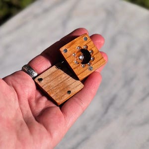 May include: Two small, square wooden boxes with a natural wood grain. One box is open, revealing a central, circular design with a small, shiny gem. The boxes are held in a hand, showcasing the craftsmanship and detail of the wooden pieces.