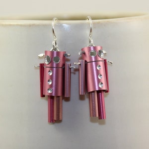 May include: A pair of pink robot earrings made from aluminum. The robots have silver accents and are hanging from silver hooks.