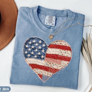 May include: A light blue t-shirt featuring a heart-shaped American flag design. The flag is made of red, white, and blue fabric patches and white stars. A gold necklace with a pendant rests on the shirt. The tag on the shirt reads "Comfort Colors".