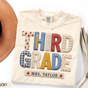 May include: An ivory t-shirt featuring the text THIRD GRADE in large, multi-patterned fabric letters, including apple prints, a pencil, plaid, and alphabet motifs. Below, a rectangular patch displays the text MRS. TAYLOR with two red apples.