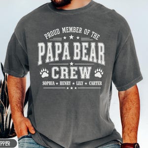 Könnte beinhalten: Ein dunkelgraues T-Shirt mit dem weißen Text "PROUD MEMBER OF THE PAPA BEAR CREW". Darunter stehen die Namen "SOPHIA * HENRY * LILY * CARTER". Das Design umfasst Pfotenabdrücke und Sterne.
