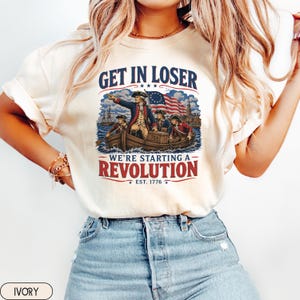Könnte beinhalten: Ein elfenbeinfarbenes T-Shirt mit einem Grafikdruck von historischen Figuren in einem Holzboot mit einer amerikanischen Flagge. Der Text lautet GET IN LOSER WE'RE STARTING A REVOLUTION EST. 1776 in blauer und roter Schrift. Kurzärmeliges T-Shirt.