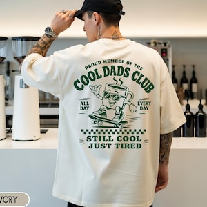 Könnte beinhalten: Ein elfenbeinfarbenes Kurzarm-T-Shirt mit einer dunkelgrünen Grafik auf der Rückseite. Das Design zeigt eine Comic-Kaffeetasse auf einem Skateboard mit dem Text "PROUD MEMBER OF THE COOL DADS CLUB" und "STILL COOL JUST TIRED" in Fettschrift.