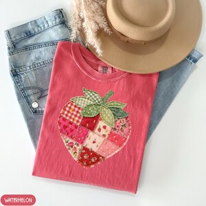 May include: A pink t-shirt featuring a patchwork strawberry design, a tan hat, and light blue jeans. The strawberry is made of various patterned fabrics, including gingham and floral prints. The word "WATERMELON" is in a red oval.