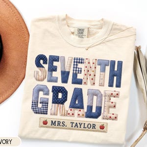 May include: An ivory t-shirt featuring the text SEVENTH GRADE in large, multi-patterned blue and beige fabric letters. Below the text is a rectangular patch with the name MRS. TAYLOR and two small red apple icons. The shirt has a crew neckline.