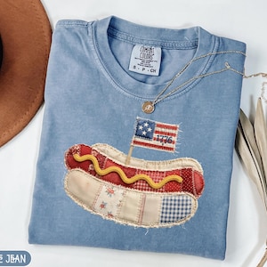 Könnte beinhalten: Hellblaues T-Shirt mit einem patriotischen Hotdog-Applikation. Der Hotdog hat ein rot-weißes Brötchen, eine Senf-Schlange und eine kleine amerikanische Flagge mit dem Jahr "1776". Eine goldene Halskette liegt über dem Shirt.