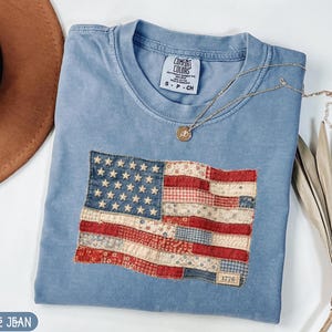 Puede incluir: Una camiseta azul claro con un diseño de bandera estadounidense cosido. La bandera presenta un diseño de patchwork con telas rojas, blancas y azules. Un collar de oro con un pequeño colgante descansa sobre la camisa. La camisa tiene una etiqueta que dice "Comfort Colors".