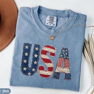 Puede incluir: Una camiseta azul claro con la palabra "USA" aplicada en la parte delantera. Las letras están hechas de tela de retazos con patrones rojos, blancos y azules. Un collar dorado con un colgante descansa sobre la camiseta.