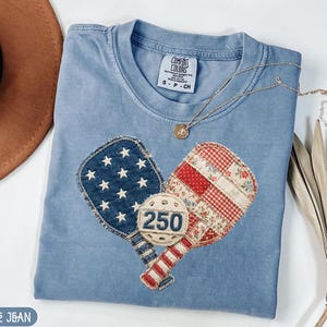 May include: A pale blue t-shirt with a heart-shaped design featuring two pickleball paddles and a ball. One paddle has a blue star pattern, the other has a red and white floral pattern. The ball has the number "250" on it.