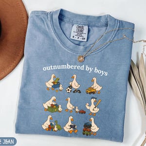 May include: A blue jean coloured t-shirt with the phrase "outnumbered by boys" in white. The shirt features cartoon ducks with toys. A gold necklace with a pendant is draped across the shirt.