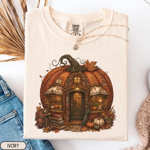 May include: An ivory t-shirt featuring a graphic print of a large orange pumpkin shaped like a cozy bookstore. The design includes a central door, windows, stacks of books, autumn leaves, and small pumpkins. The shirt has a Comfort Colors brand label.
