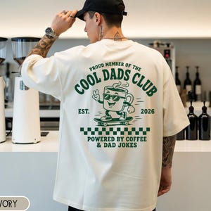 May include: Ivory-colored t-shirt with a green graphic that reads "Proud Member of the Cool Dads Club" and a cartoon coffee cup on a skateboard. The text "Powered by Coffee & Dad Jokes" is also featured. The shirt has a classic fit.