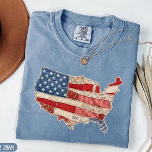 Puede incluir: Una camiseta Comfort Colors azul claro con un diseño de bandera estadounidense cosido en forma de Estados Unidos. La bandera está hecha de varias telas estampadas. Un collar de oro con un colgante descansa sobre la camisa.