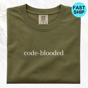 May include: An olive green crew neck t-shirt featuring the text code-blooded printed in white across the center chest. The shirt has a Comfort Colors brand label at the inner neckline indicating a size large, with a ribbed collar and short sleeves.