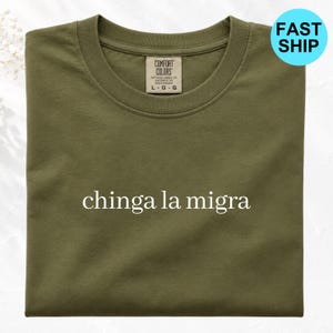 以下が含まれることがあります： 胸の中央に白い文字でchinga la migraとプリントされたオリーブグリーンのクルーネックTシャツ。衣類の内側のネックラインにはComfort Colorsのブランドラベルがあり、サイズはラージでホンジュラス製であることが示されています。