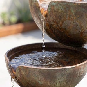 Peut inclure: Fontaine d'extérieur à trois niveaux avec des bassins en forme de bol. L'eau coule du bassin supérieur, créant une douce cascade. La fontaine a une finition patinée, semblable à du bronze, ajoutant une esthétique naturelle au jardin.