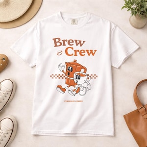Puede incluir: Camiseta blanca con un diseño retro de una cafetera y una taza de café caminando juntas. El texto "Brew Crew" y "Fueled by Coffee" está impreso en marrón. Es una prenda de vestir informal.