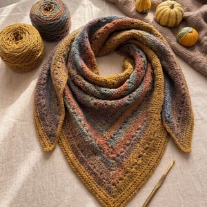 Pode incluir: Um xale de crochê multicolorido feito à mão em tons de dourado, castanho, azul e pêssego. O xale é triangular, com um padrão de ponto texturizado. Duas bolas de lã e uma agulha de crochê estão ao lado do xale. O tema é aconchegante e artesanal.