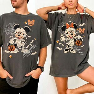 May include: A charcoal gray t-shirt featuring a graphic of a cartoon mouse wrapped in bandages like a mummy, holding a jack-o'-lantern. The design includes a spiderweb, bats, candy corn, and the text "Minnie Mouse" in a cursive script.