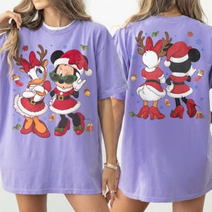 Pode incluir: Uma t-shirt roxo-claro com uma impressão gráfica da Daisy Duck e da Minnie Mouse vestidas com trajes de Natal vermelhos e brancos. A frente mostra-as de frente, enquanto as costas as mostram de trás, rodeadas por flocos de neve.