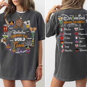 May include: A charcoal gray short-sleeve t-shirt featuring a front graphic with the text Drinking Around The World Team surrounded by various drink icons and spiderwebs. The back displays the text Drinking Around The World with country flags.