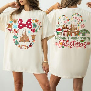 May include: An off-white t-shirt featuring a front graphic of a gingerbread castle, a red polka-dot bow, and holiday icons. The back displays a gingerbread castle, a globe, a tree, and the text Mickey's very merry Christmas 2024.