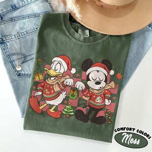 May include: A moss green crewneck t-shirt featuring a graphic of Donald Duck and Mickey Mouse wearing Santa hats and Christmas sweaters. The design includes a red and green checkered background with holiday icons like a tree and wreath.