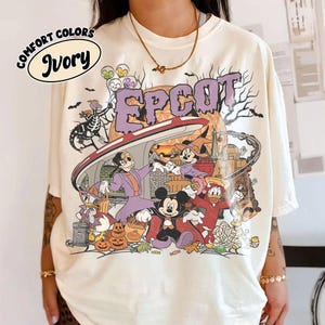 May include: An ivory t-shirt featuring a graphic print with the text EPCOT in purple letters. The design includes cartoon characters dressed in Halloween costumes, a monorail, pumpkins, and a skeleton figure against a backdrop of trees.