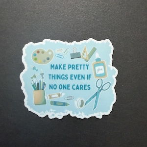 May include: A light blue sticker with the text "MAKE PRETTY THINGS EVEN IF NO ONE CARES." The sticker features illustrations of art supplies, including a paint palette, scissors, glue, and pencils.