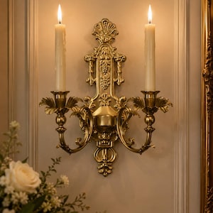 May include: A gold-toned metal wall sconce featuring two candle holders with lit off-white taper candles. The sconce has an ornate, symmetrical design with leaf-like accents and a central vertical panel decorated with embossed grape clusters.