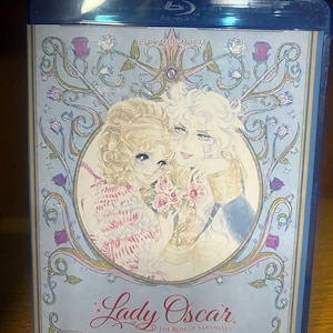 May include: A blue Blu-ray case with the title "Lady Oscar" and an illustration of two figures embracing, surrounded by floral and ornate designs. The case includes Korean text and a "Blu-ray Disc" logo.