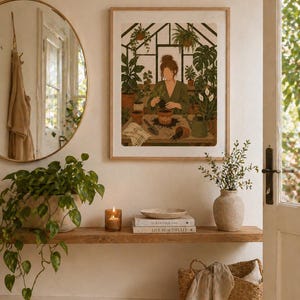 Op de afbeelding: Een ingelijste kunstafdruk toont een persoon die planten verzorgt in een kas. Het kunstwerk bevindt zich in een houten lijst boven een houten plank. De plank toont boeken, een kaars en een vaas met groen. Een ronde spiegel en een rieten mand zijn ook zichtbaar.