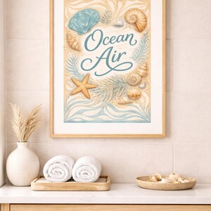 Op de afbeelding: Een ingelijste print met de tekst "Ocean Air" in een cursief lettertype, omringd door illustraties van schelpen en een zeester. Het kunstwerk wordt weergegeven boven een houten wastafel met opgerolde witte handdoeken en decoratieve items.